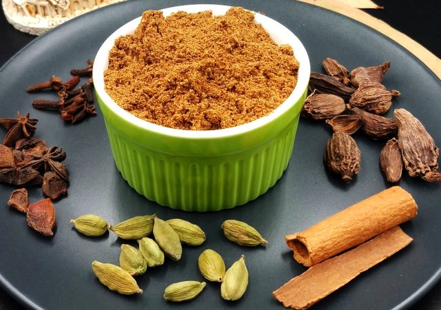 Ground biryani masala powder in green small bowl on black plate with some gram masala