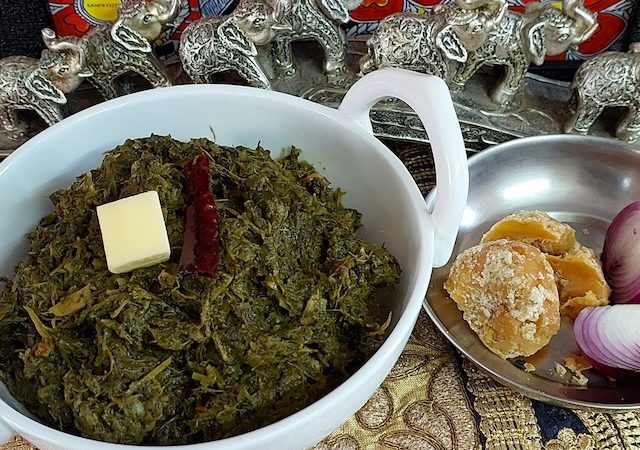 A bowl of Sarson ka Saag served with a piece of jaggery and onion, showcasing the vibrant green colors of the dish.