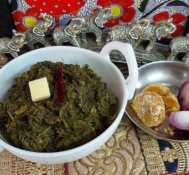 A bowl of Sarson ka Saag served with a piece of jaggery and onion, showcasing the vibrant green colors of the dish.

