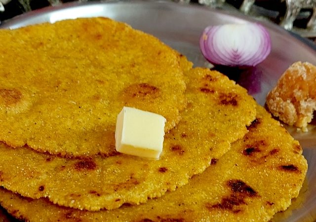 Image of freshly cooked Makki Ki Roti on a hot griddle, with golden-brown spots, served with a side of jaggery and onion piece.